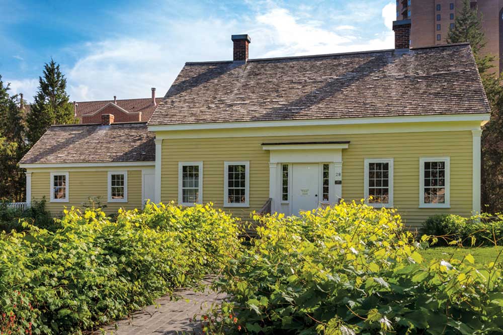 The frontside of the Ard Godfrey house. A charming home that is painted yellow with bushes lining the walkway up to the front door.
