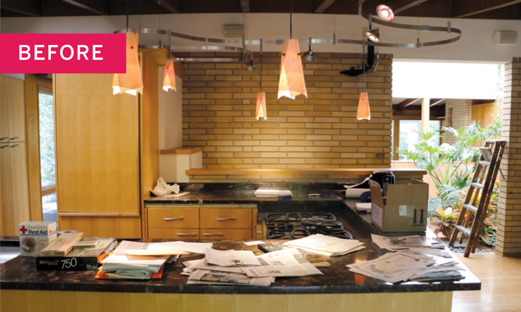 A cramped and cluttered kitchen lightly-colored wood cabinetry, and granite countertops with papers scattered across.