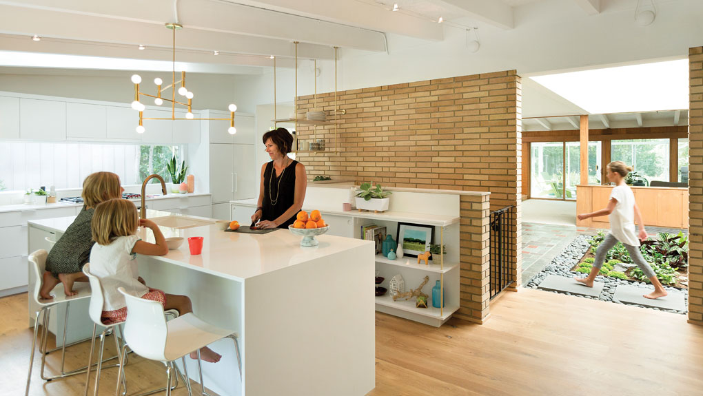 The owner of the home prepares a snack for her daughters as they wait at the counter. A third daughter is walking in the indoor atrium in the background.
