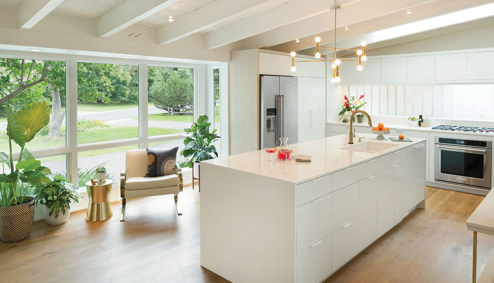 A sleek, updated white kitchen with large windows flanked by plants looks out at the driveway and green surroundings.
