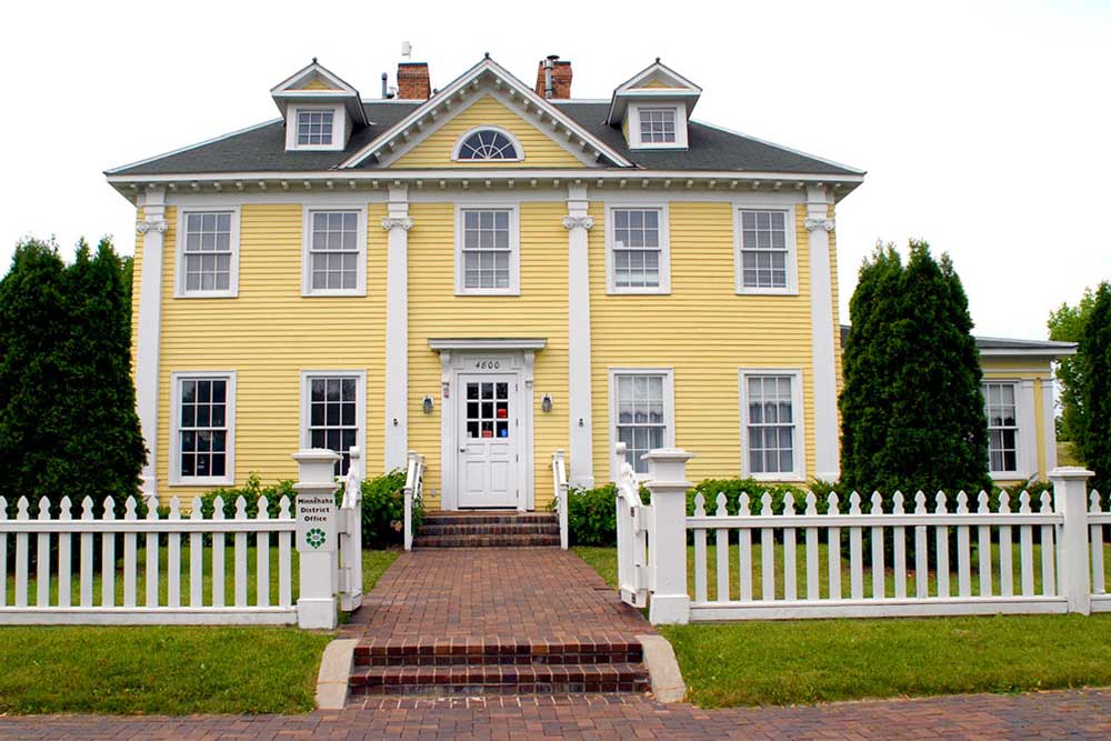 The frontside of a Longfellow House shows a white picket fence outside of a large, two-story, symmetrical yellow home. A brick path leads up to the white front door. Each side of the home features two windows on each story.
