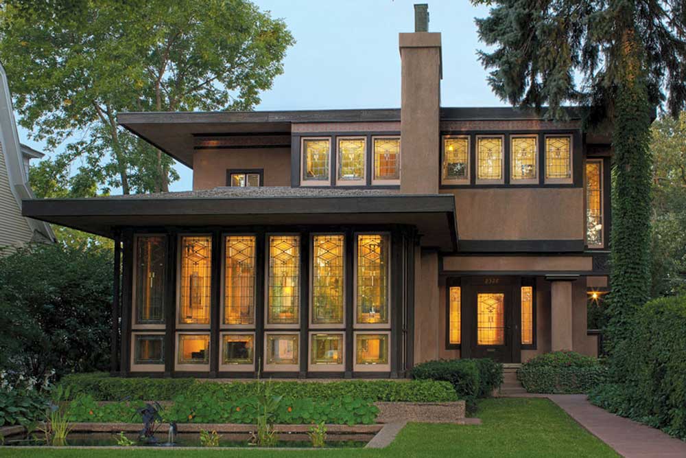 The Purcell-Cuts House. In the front yard is a small pond with a small fountain in the center. The home features two stories with floor-to-ceiling windows and flat roofs. Plants line the home and wrap around back.