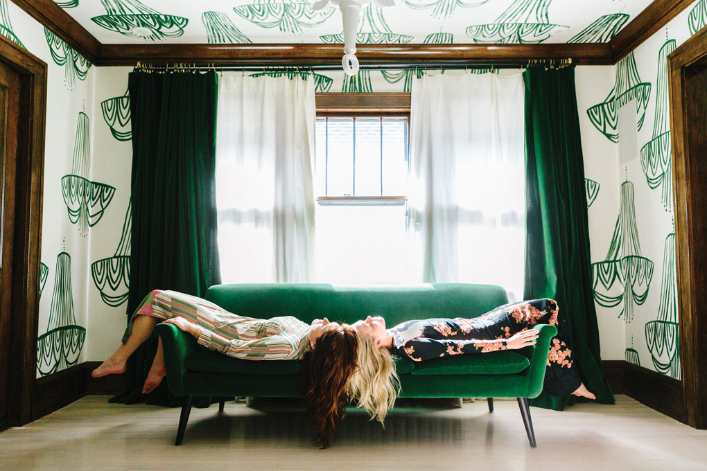 Jennifer Jorgensen and Kate Worum of She She lie on a green couch in a room covered in wallpaper featuring hand drawn chandeliers.