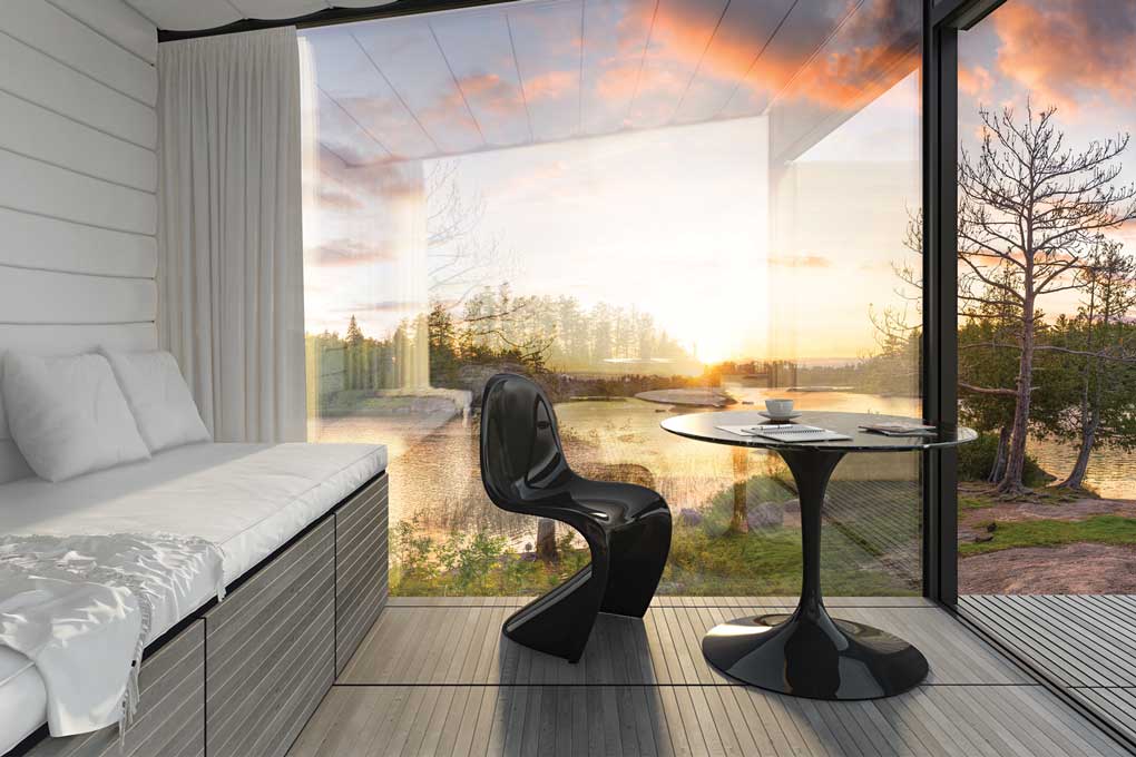 A sun room with walls made of glass looks out onto a lake surrounded by a forest as the sun sets.