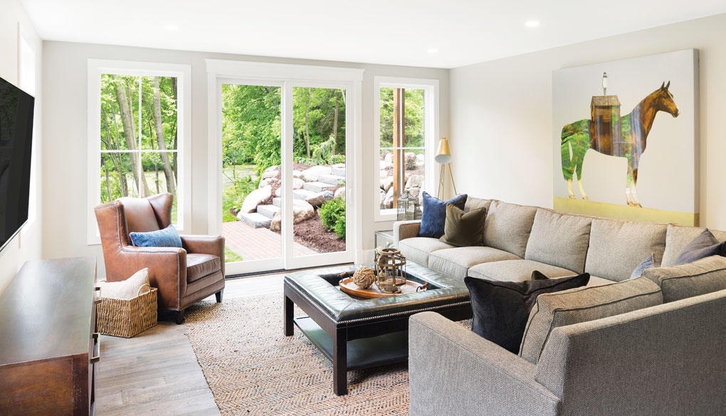 A basement furnished with sofas and chairs sits facing a wall with a TV mounted onto it. On the opposite wall is a horse painting. A walkway leads out to the backyard.
