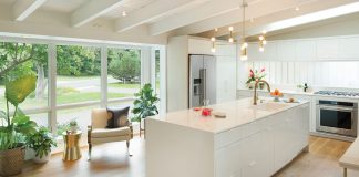 A sleek, updated white kitchen with large windows flanked by plants looks out at the driveway and green surroundings.