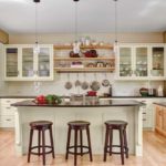 Digiacomo Homes, Inc. A kitchen designed by Gigi Digiacomo of Digiacomo Homes, Inc. shows light-colored hardwood flooring, white cabinetry, black countertops and an island surrounded by dark-colored wooden stools.
