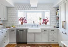 A kitchen designed by Kate Roos of Kate Roos Design, LLC features all white cabinetry and countertops with a white subway tiled backsplash. In the large window over the sink sit two pink flowers.