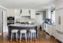 A kitchen designed by Liz Schupanitz Designs on the 2017 ASID Designer Kitchen Tour that features white cabinetry and hardwood floors. In the center is a blue-colored island with stools surrounding it.