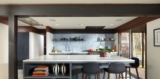 A Midcentury Modern Revival for a 1960s Home A modern kitchen features a central island with highchairs on one side of it. It also features white countertops, shelves mounted on the ceiling-high backsplash, and a range hood tucked into the ceiling.