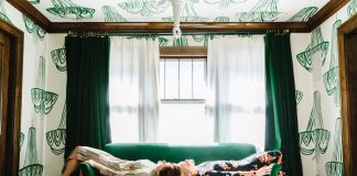 Kate Worum and Jennifer Jorgensen’s She She Offers Custom, Hand-Painted Wallpaper Jennifer Jorgensen and Kate Worum of She She lie on a green couch in a room covered in wallpaper featuring hand drawn chandeliers.