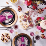 A table decorated with holiday gift ideas, including black plates and flatware, purple salad plates, deep-cut crystal goblets, white marble cheese plates, candles, flowers and more.