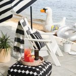 A white deck chair with a tray of beverages and umbrella next to water.
