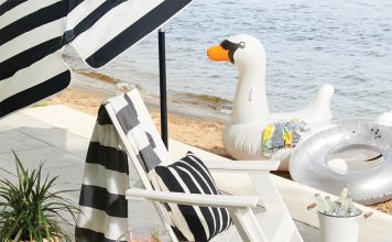 A white deck chair with a tray of beverages and umbrella next to water.