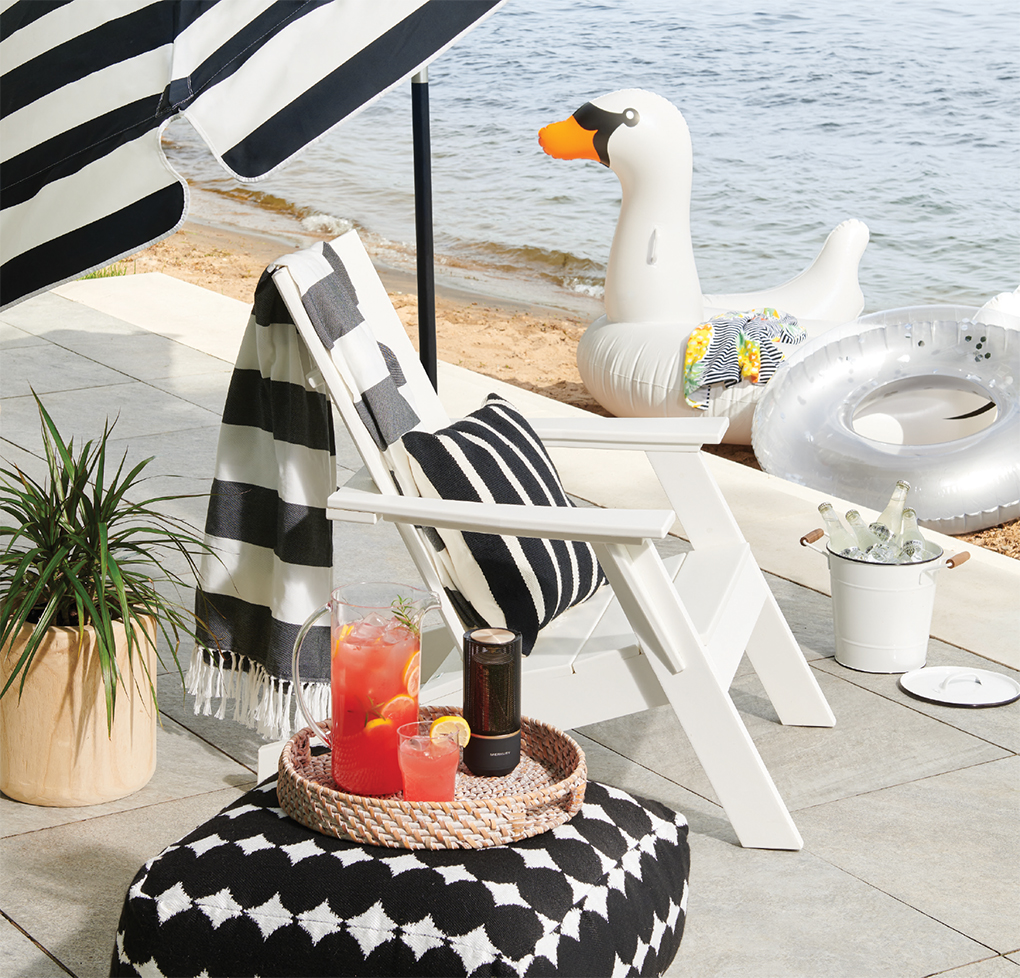An outdoor accessory picture featuring a white deck chair with a tray of beverages and umbrella next to water.