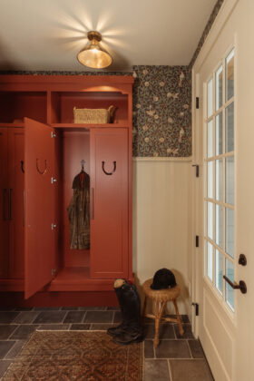 Botanical-patterned William Morris wallpaper, buttercream wainscoting, and horseshoe carvings in the coat cabinet doors add whimsy to the well-traveled mudroom.