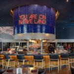 Design-Forward Twin Cities Restaurants Bring Dining to New Heights IndÃgena by Owamni interior with "You Are On Native Land" sign above the central bar. August 19, 2026