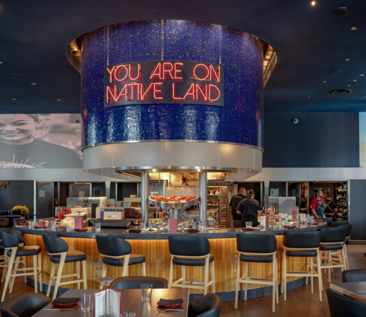 Indígena by Owamni interior with "You Are On Native Land" sign above the central bar. August 19, 2026