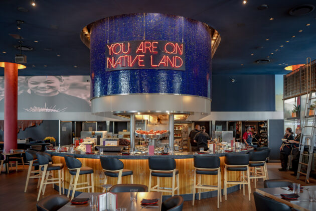 Ind&iacute;gena by Owamni interior with "You Are On Native Land" sign above the central bar. August 19, 2026