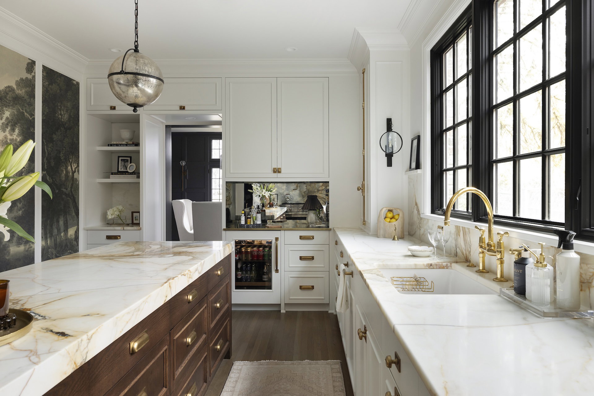 The kitchen features light cabinetry, rich brass tones, Borghini Marble countertops, a unique wall mural, a large island, and creative storage solutions.
